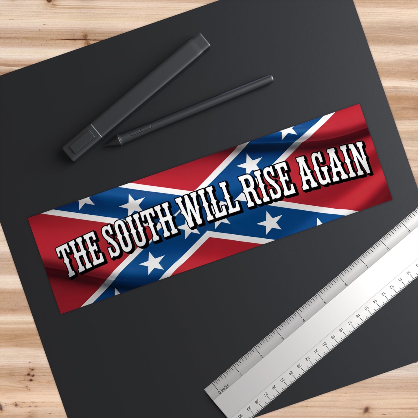 Confederate flag bumper sticker with the text The South Will Rise Again on black surface with pen and ruler