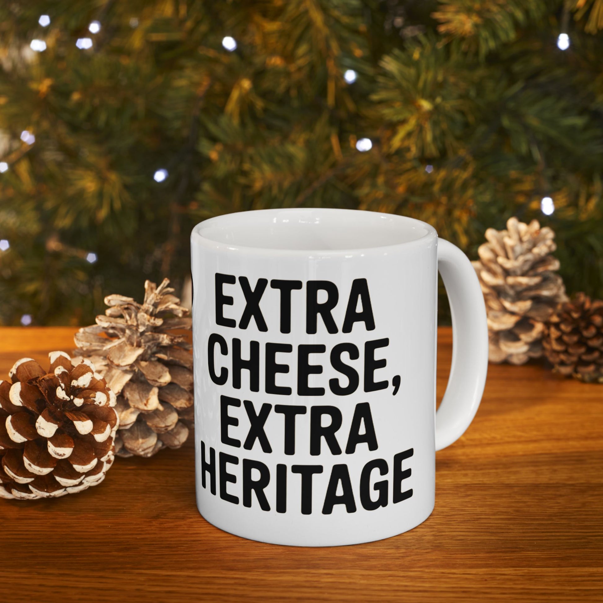 White mug with black text reading Extra Cheese Extra Heritage on wooden table with pine cones